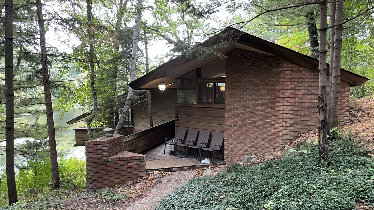 Low brick-and-cedar midcentury house with a long overhanging roof and a wooded entry walk beside a lake in Burnsville