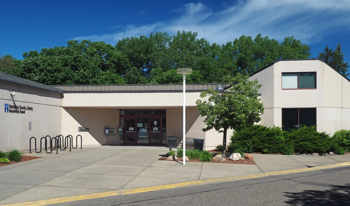 Low light-colored Rockford Road Library building with a recessed entrance, bicycle racks and mature trees in Crystal