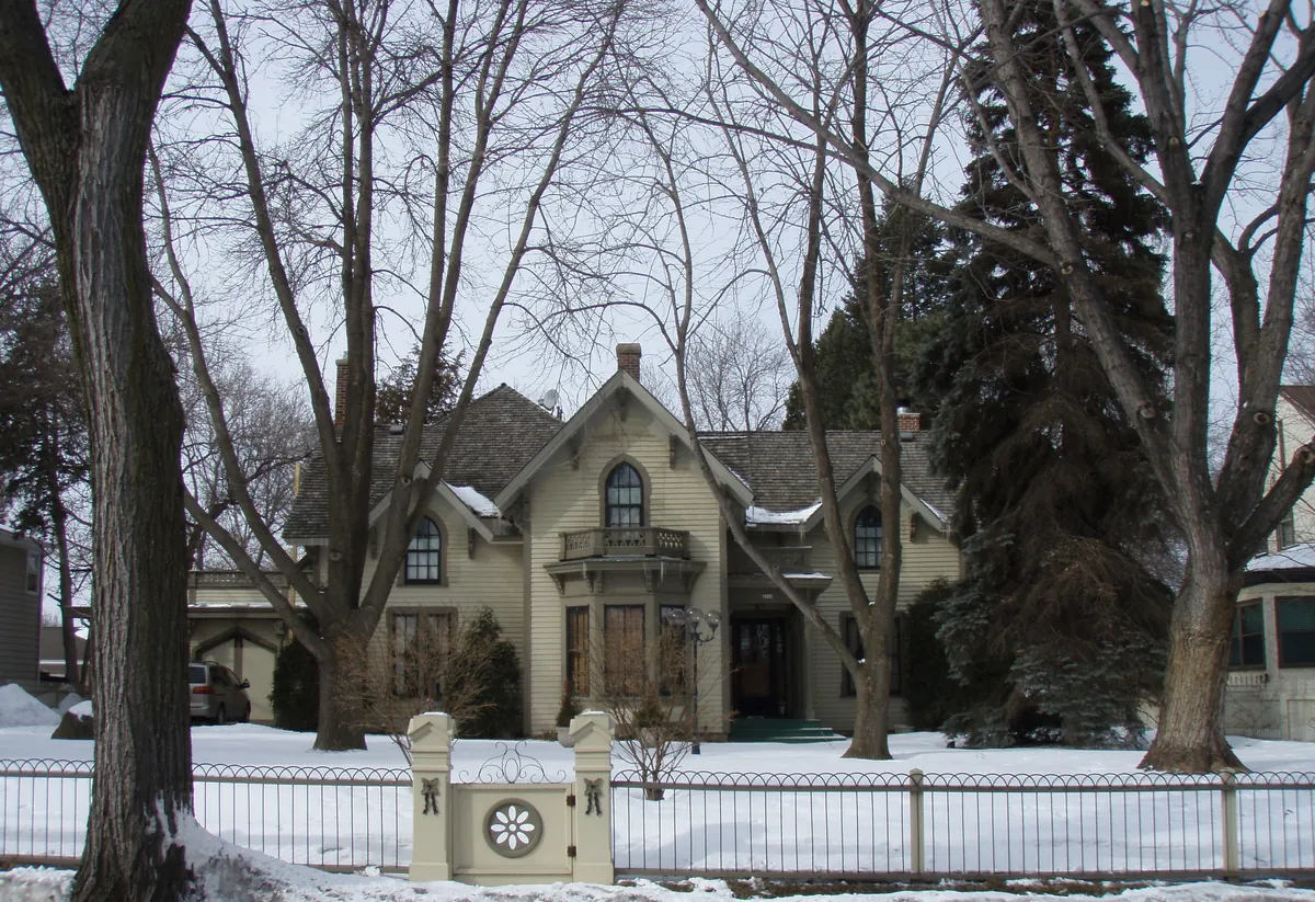 Cream Gothic Revival house with pointed-arch windows and decorative gables behind a low iron fence and mature trees in winter, Morningside, Edina
