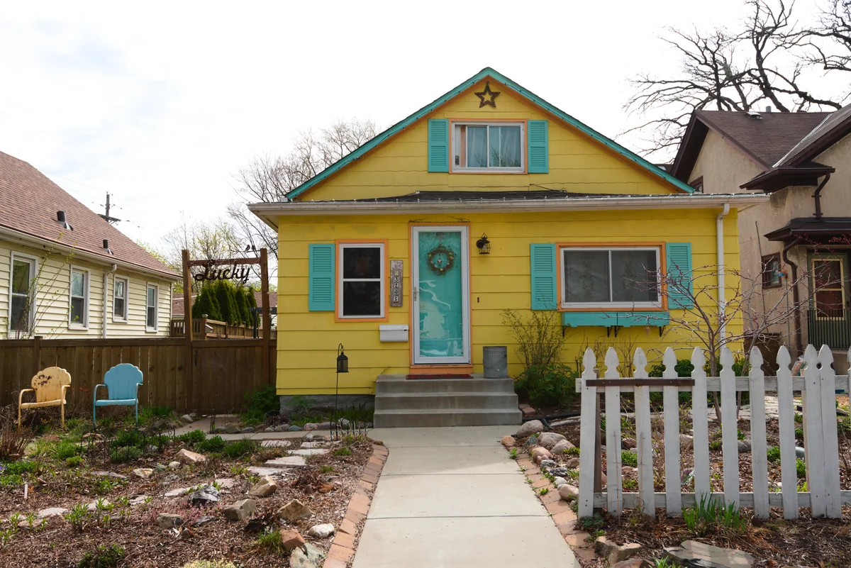 Yellow story-and-a-half bungalow with teal shutters and door, a front garden and a white picket fence in Minneapolis's Cooper neighborhood