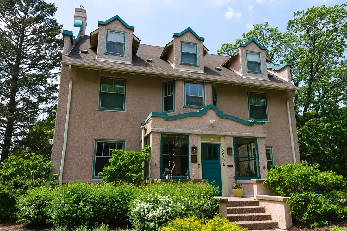 Three-story pink stucco house with teal trim, three front dormers and a curved entry parapet, framed by shrubs and mature trees near Lake Harriet