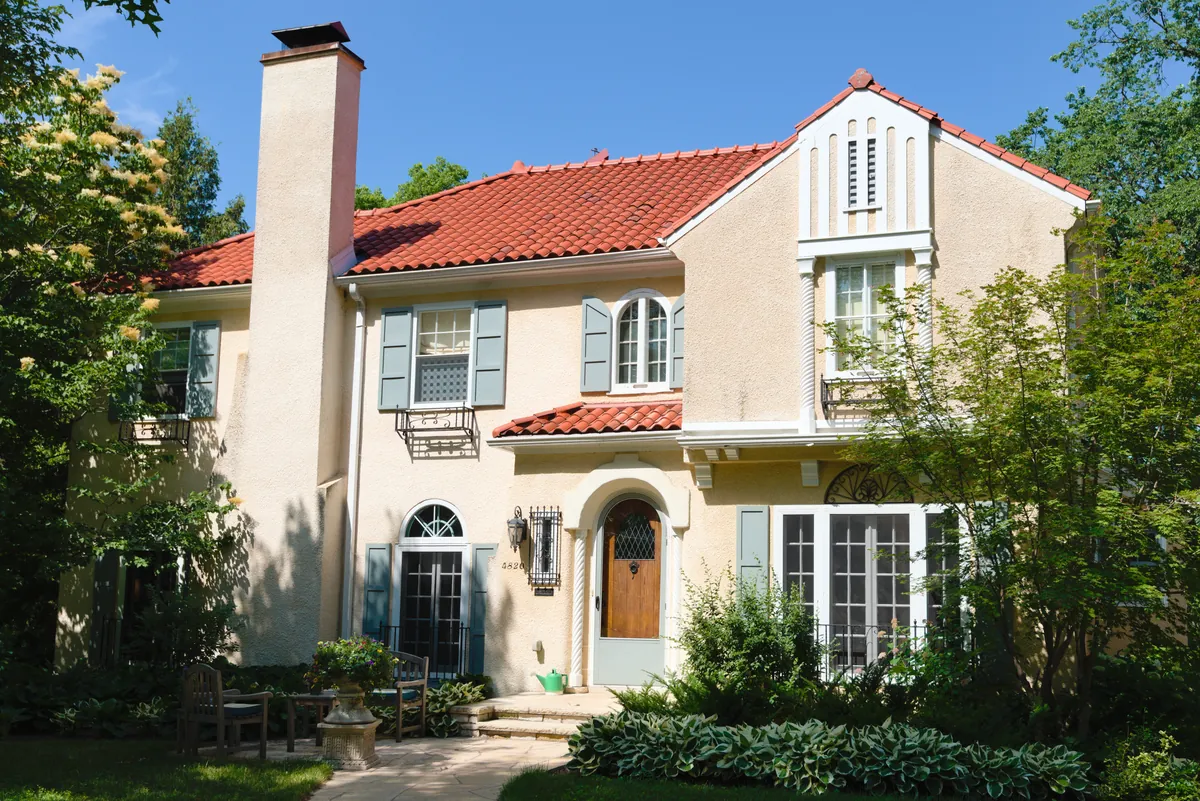 Two-story stucco house with a red clay-tile roof, gray-green shutters and an arched wooden front door, surrounded by garden plantings in Minneapolis's Lynnhurst neighborhood