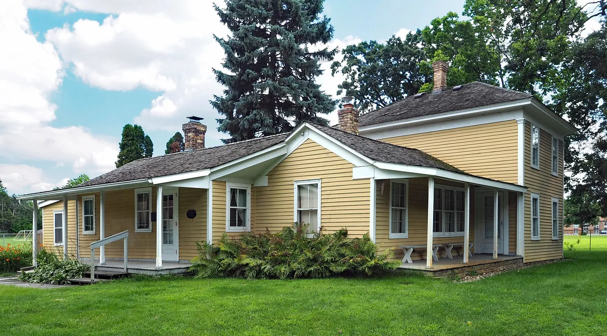 Yellow clapboard 1850s farmhouse with white trim, a long front porch and a tall spruce at the Riley Lucas Bartholomew House in Richfield