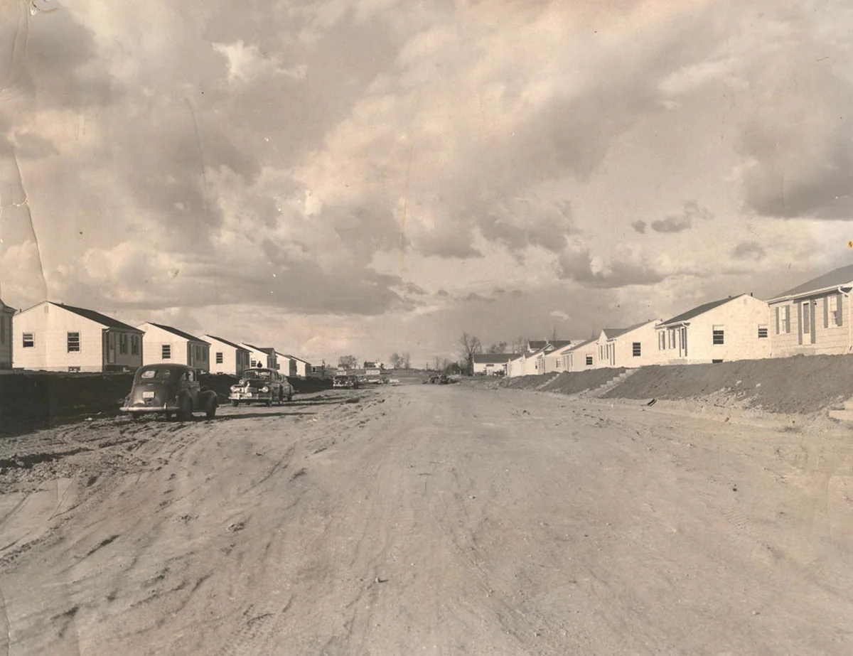 Black-and-white photo of a row of new one-story houses with lap siding and shuttered windows along unpaved Washburn Avenue in Richfield, with two 1940s cars parked on the dirt road