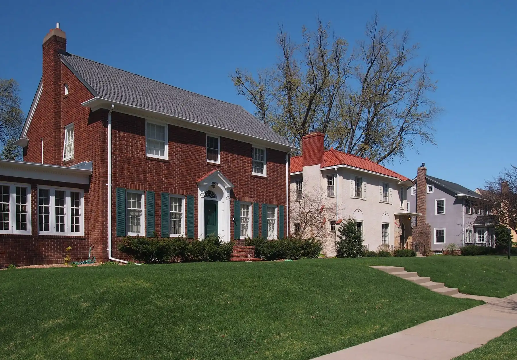 Red-brick Colonial Revival house with green shutters beside white stucco and gray homes along a sloped lawn on Bruce Avenue in Edina's Country Club district