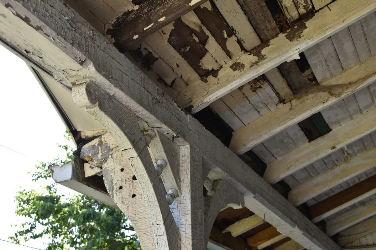 Looking up at the porch roof brackets and beadboard ceiling of an older house, where several layers of white and cream paint are cracked, flaking and worn through to bare wood