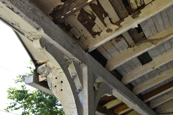 Looking up at the porch roof brackets and beadboard ceiling of an older house, where several layers of white and cream paint are cracked, flaking and worn through to bare wood