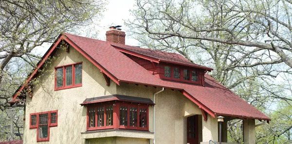A 1910 Arts and Crafts bungalow in north Minneapolis with tan stucco walls, a red shingle roof, exposed rafter tails, red painted window sash and trim, a shed dormer and an arched front porch