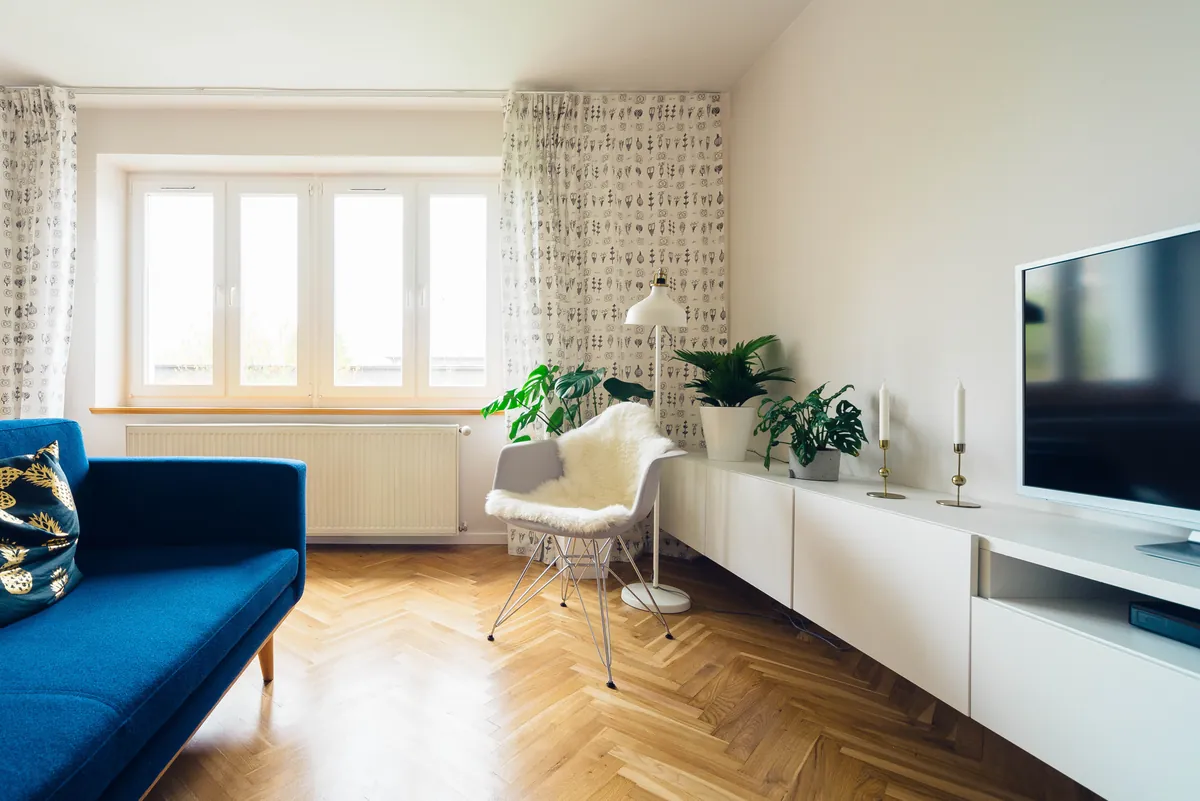 Bright living room with smooth white walls lit by daylight from a wide window, a blue sofa, a white chair, plants and a long white media cabinet on a herringbone wood floor