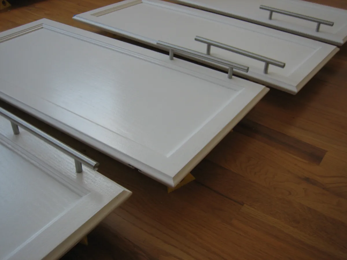 Freshly painted white raised panel cabinet doors with brushed steel bar pulls resting on painter's pyramids on a hardwood floor indoors, spaced apart