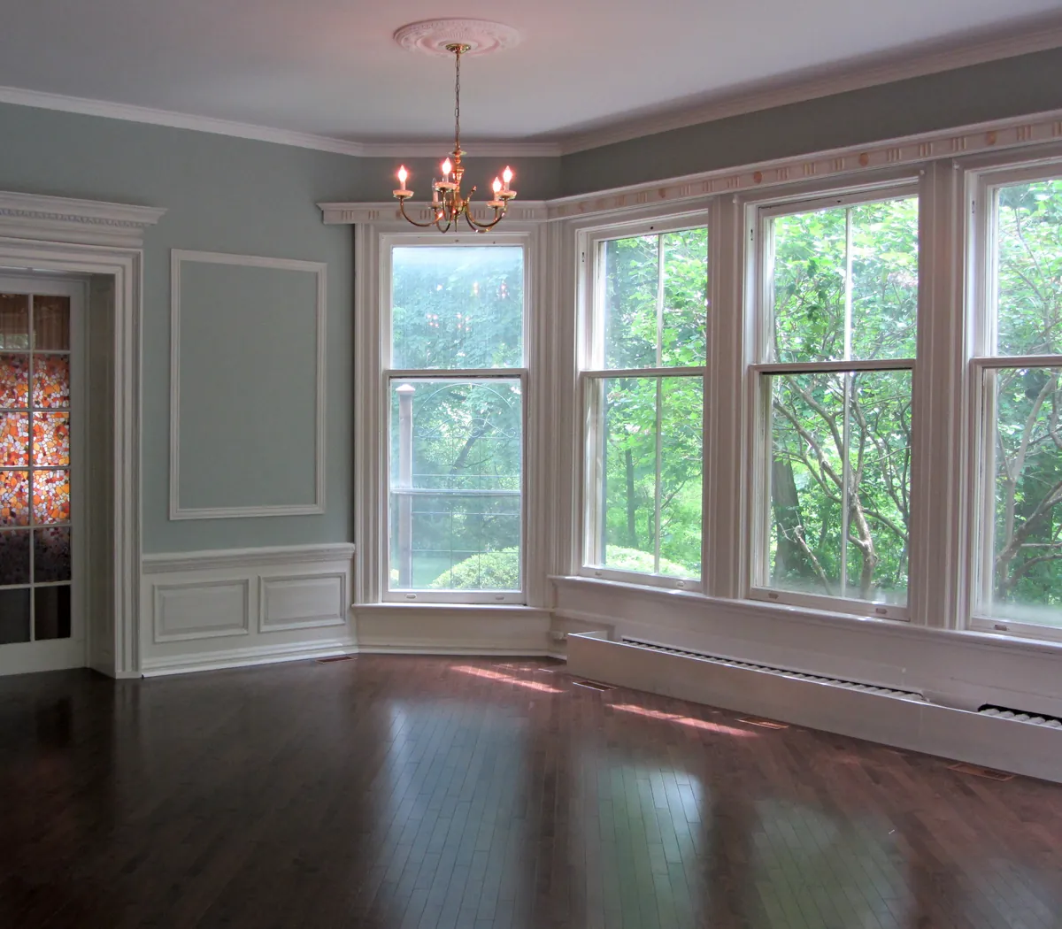 Empty room with pale blue gray walls, white crown molding, wainscoting and window trim, a bay of tall windows and dark hardwood floors