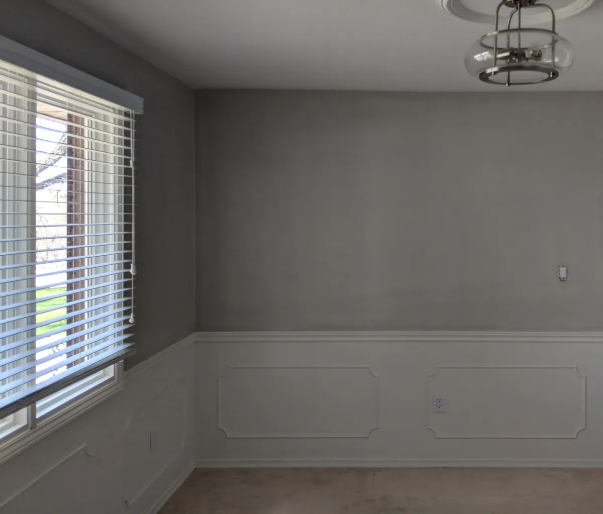 Empty dining room with a freshly painted gray wall above white wainscoting, lit from the side by a window with blinds