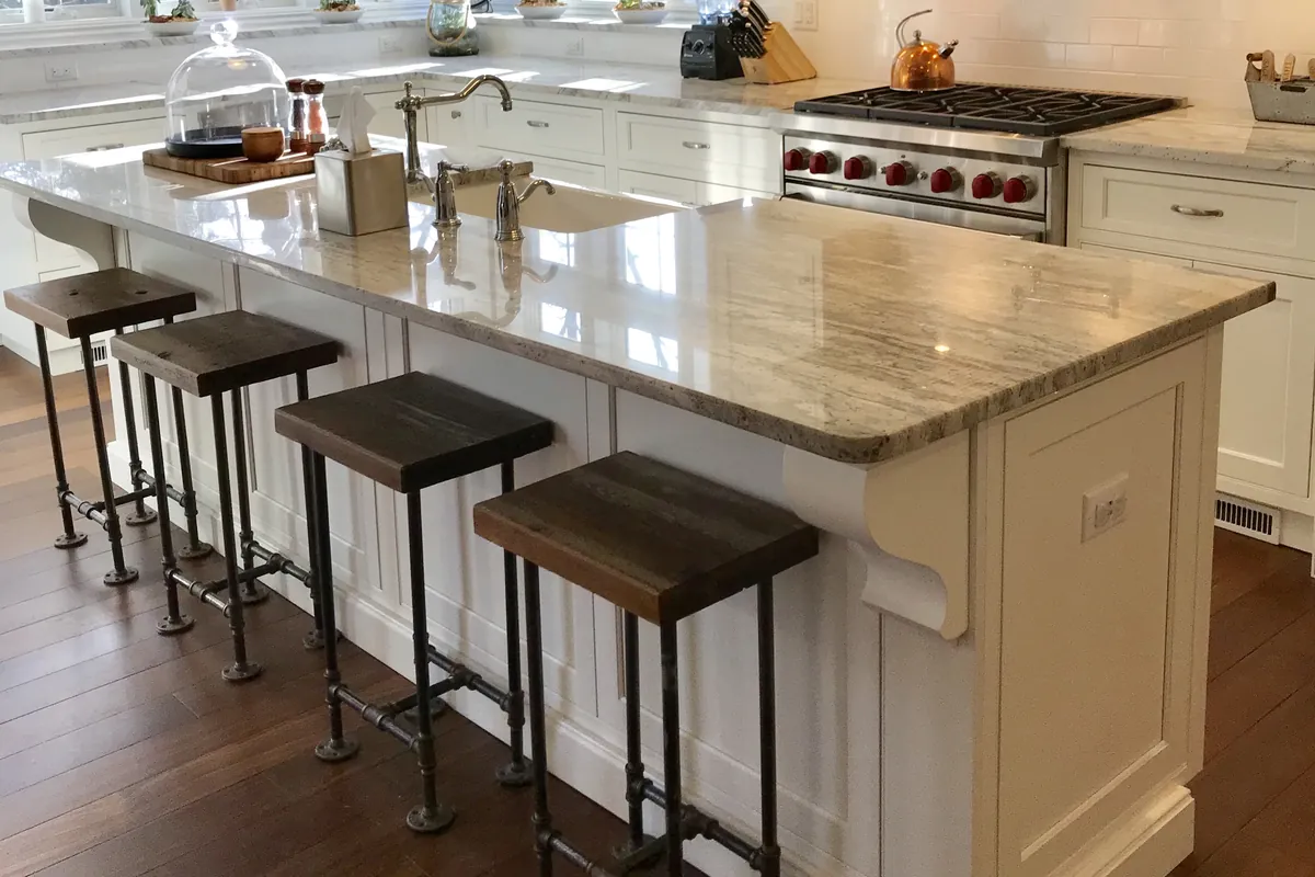 Finished kitchen with white painted cabinets, a large white island with a granite top and a corbel under the overhang, wood and pipe bar stools and a stainless range