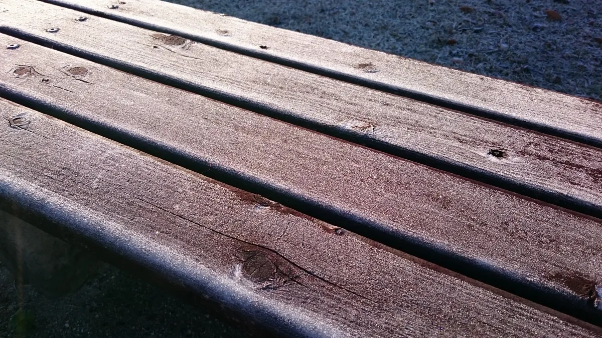 White frost crystals coating the stained wooden slats of an outdoor bench, with frosted grass behind it in early morning light