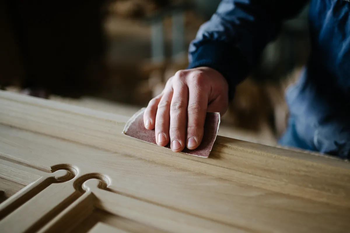 Close view of a hand pressing a folded sheet of sandpaper along the frame of a raised panel oak door laid flat