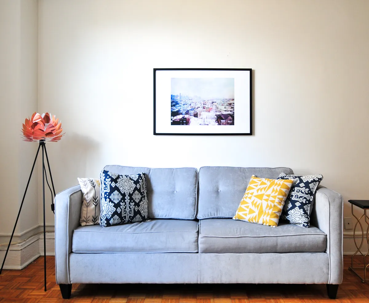 Cream painted living room wall with a framed print above a light gray sofa with patterned pillows, a copper floor lamp at left and white baseboard trim on a parquet floor