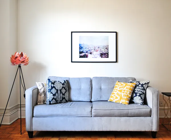 Cream painted living room wall with a framed print above a light gray sofa with patterned pillows, a copper floor lamp at left and white baseboard trim on a parquet floor