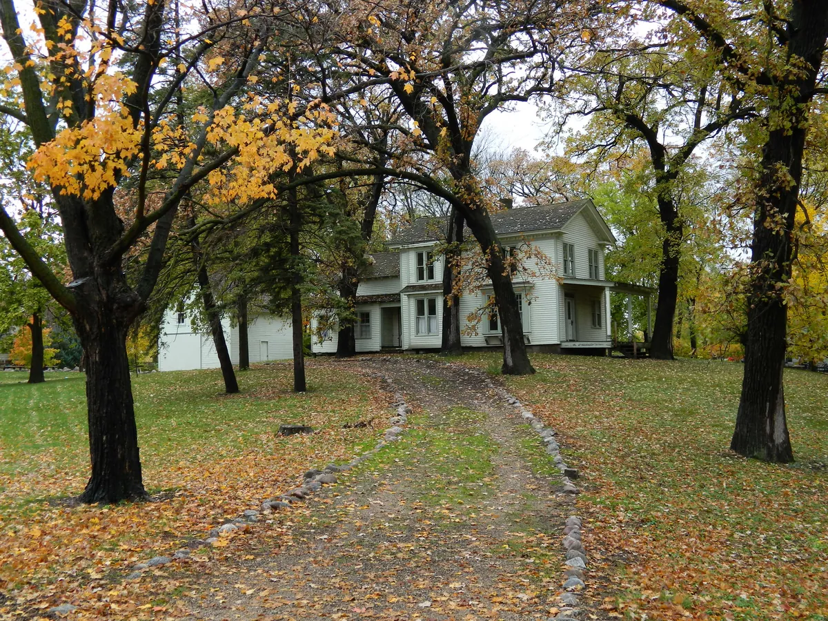 White two-story clapboard farmhouse among oak and maple trees with yellow fall leaves, a leaf-covered lawn and a stone-edged gravel drive in late October in Hutchinson, Minnesota