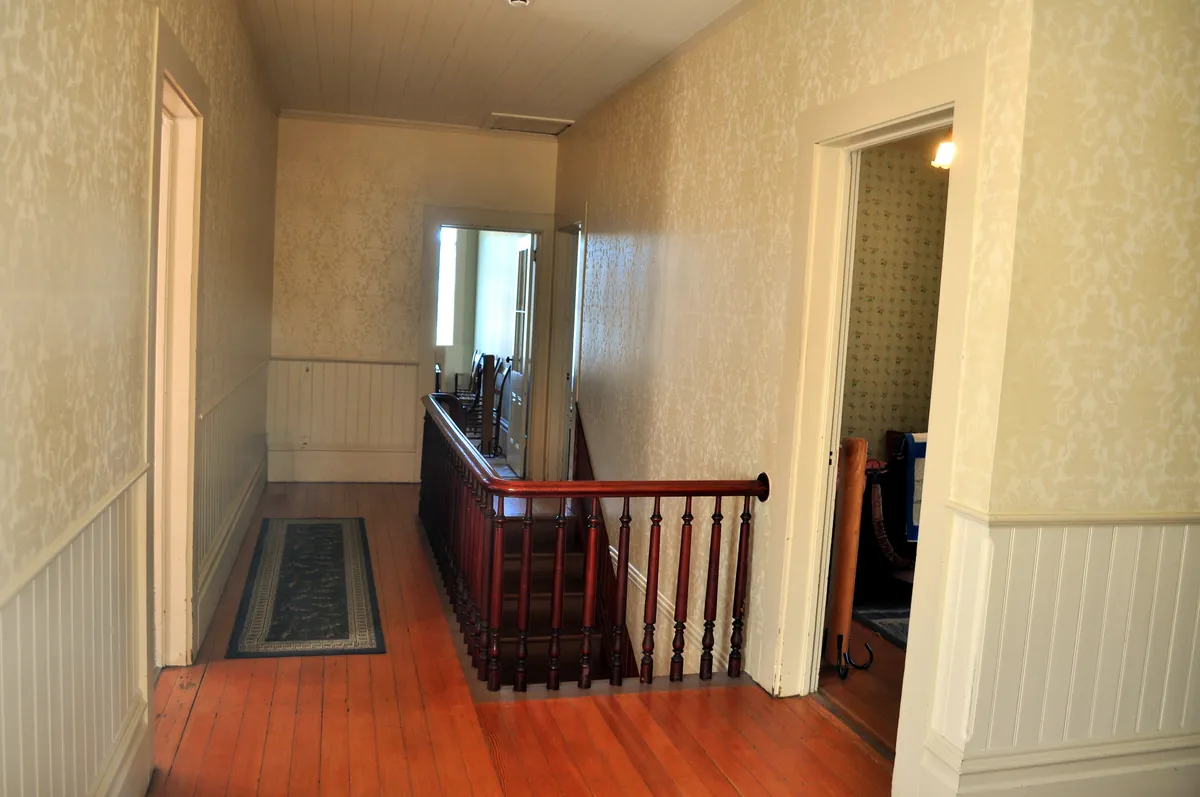 Upstairs hallway of an older house with painted cream door casings, white painted beadboard wainscoting, patterned wallpaper, a wood stair railing and fir plank floors