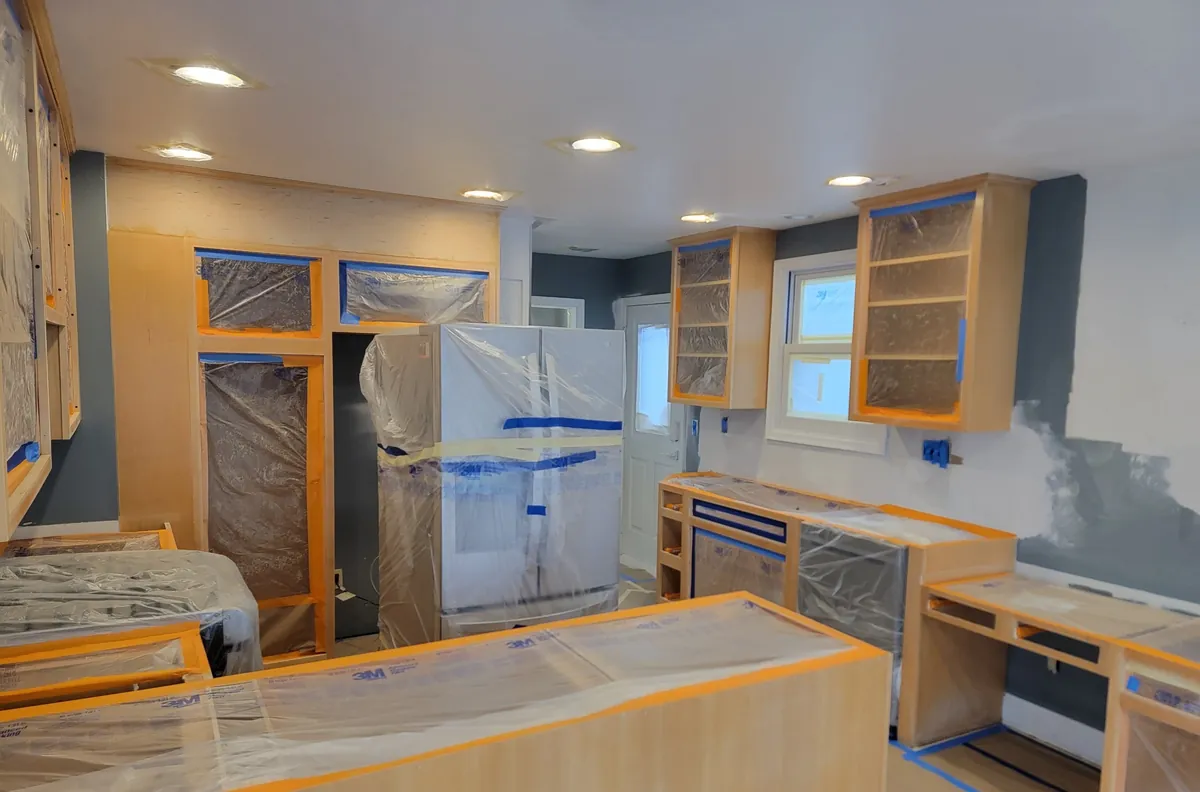 A kitchen mid-project with every cabinet door and drawer front removed, boxes and counters masked in plastic and tape, and the refrigerator wrapped
