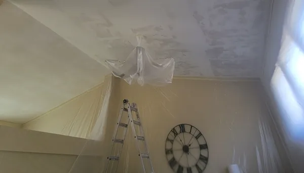 A room sealed for ceiling work: walls and windows covered in plastic, the light fixture bagged, a ladder in place and fresh skim coat on the ceiling