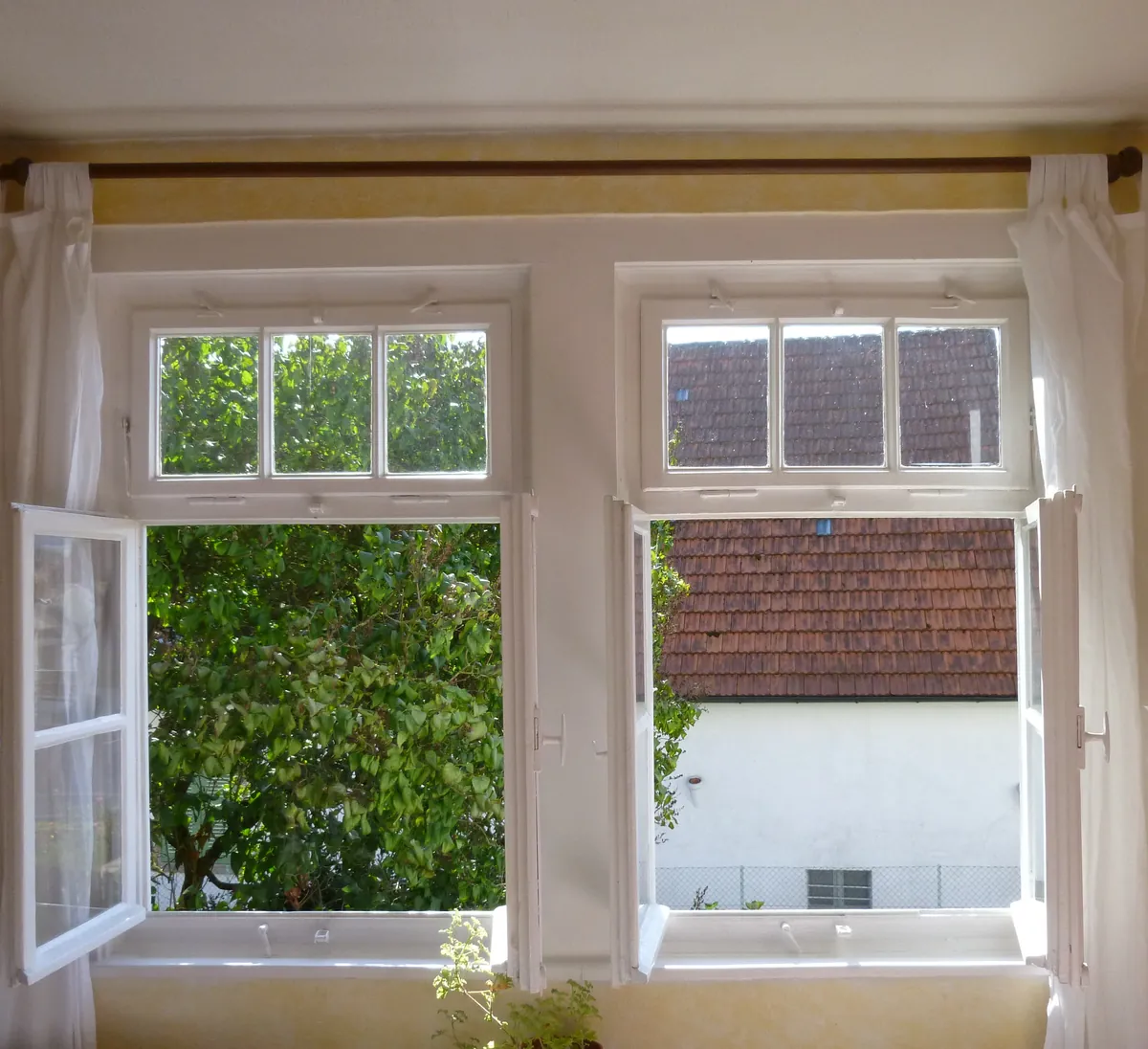A sunny yellow painted room with two tall windows swung wide open to green trees and a neighboring roof, white curtains pulled back and a small potted plant on the sill