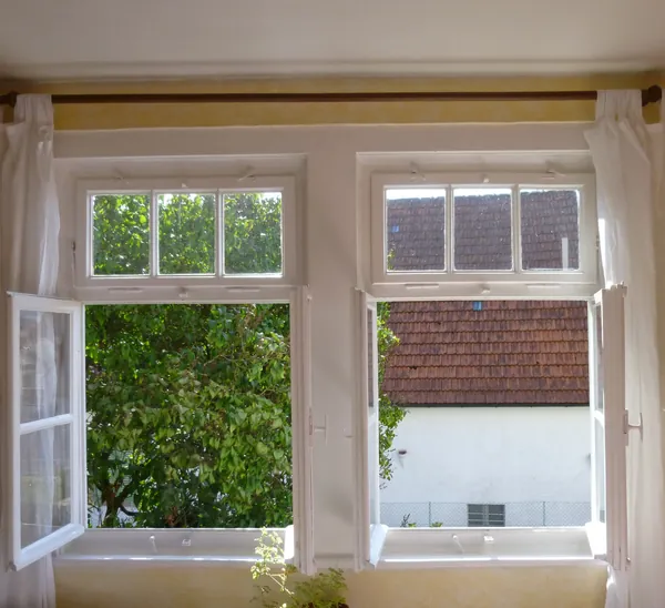 A sunny yellow painted room with two tall windows swung wide open to green trees and a neighboring roof, white curtains pulled back and a small potted plant on the sill