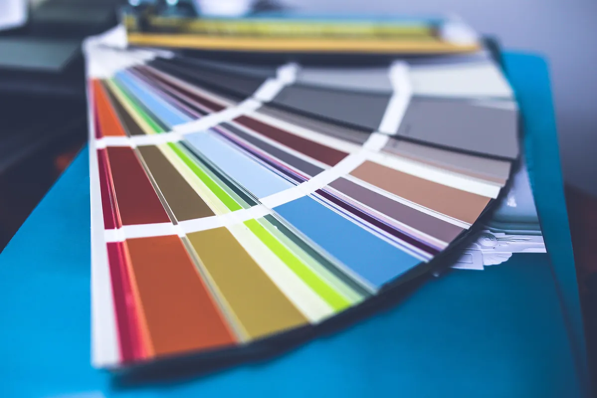 Paint color fan deck spread open on a blue folder, showing strips of red, orange, gold, green, blue, gray and brown samples with a shallow depth of field