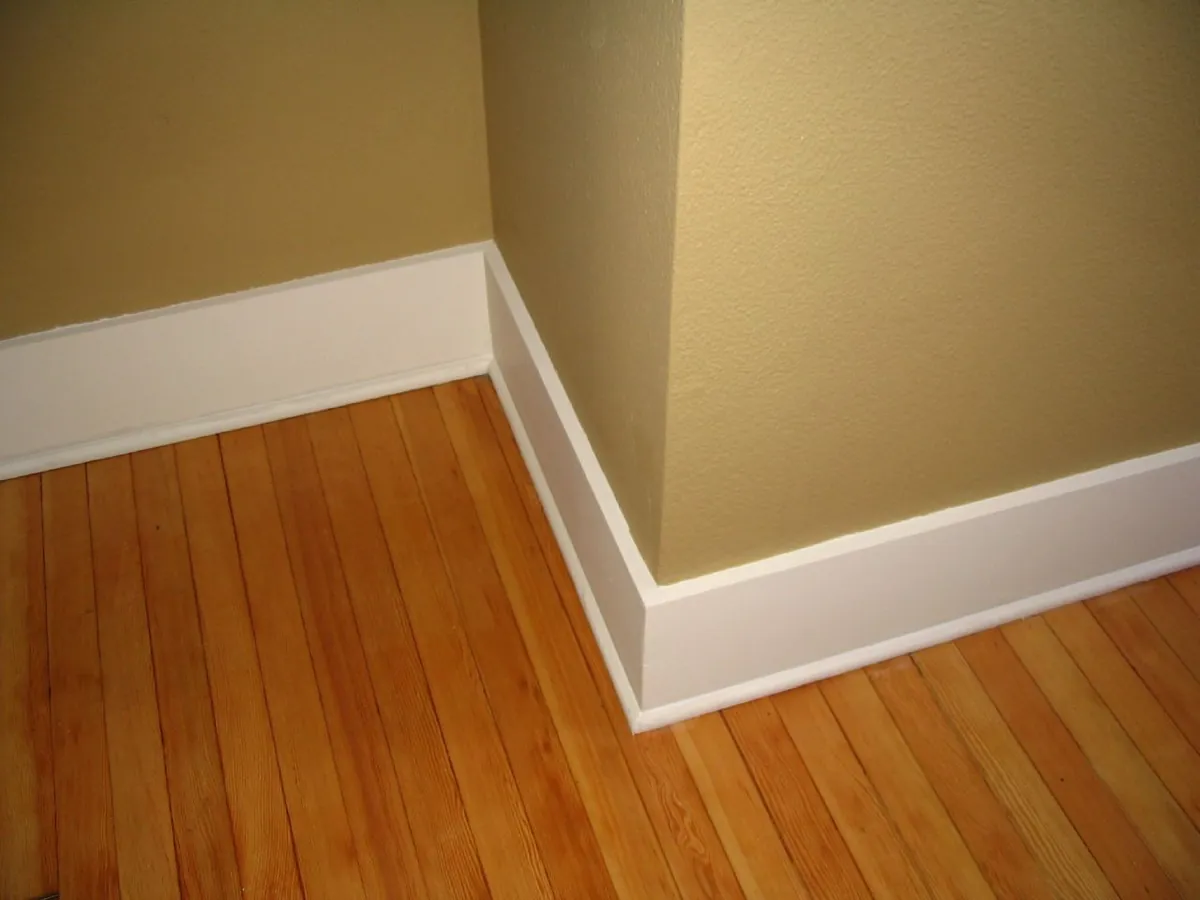 Freshly painted white baseboard and base shoe molding wrapping an outside corner against a tan wall above a honey colored wood floor