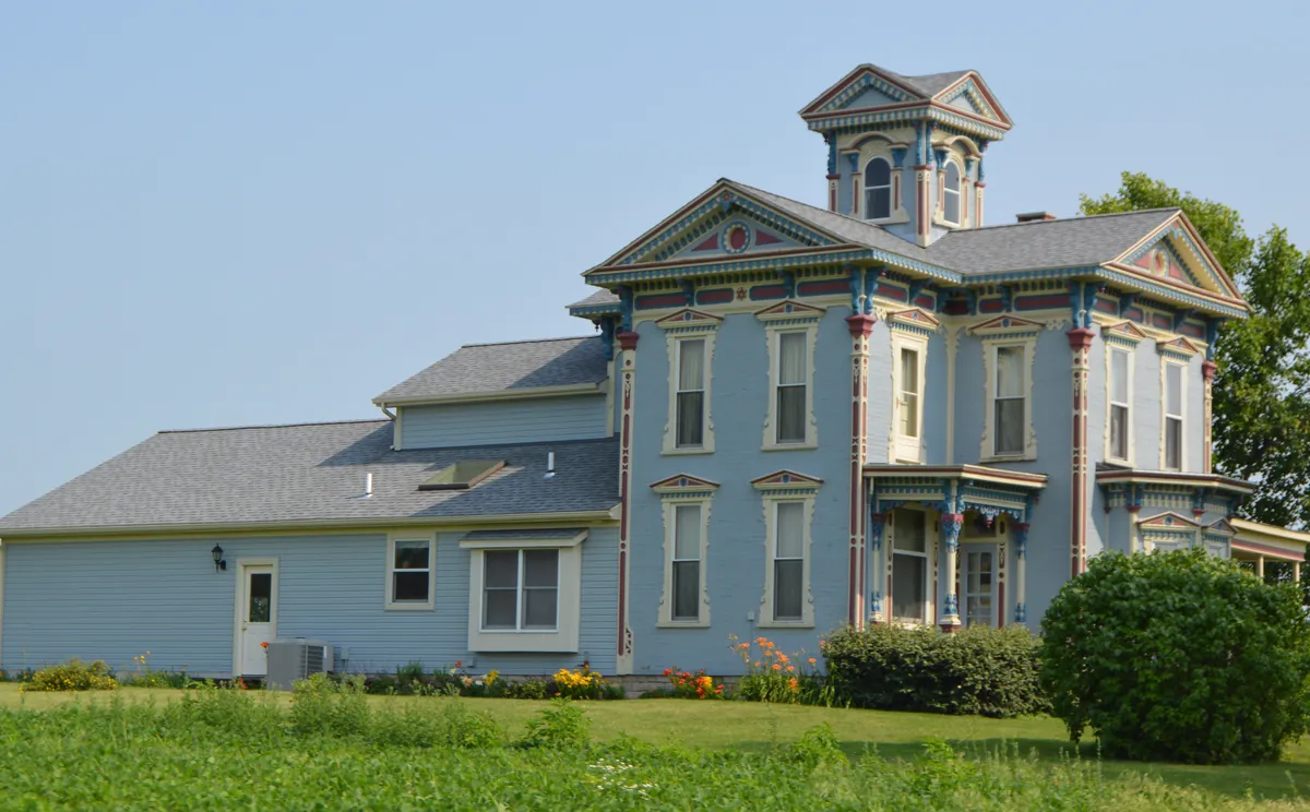 Two-story Italianate farmhouse painted light blue with cream window trim and red and blue accent colors on the brackets, cupola and porch, on a green lawn under a clear summer sky