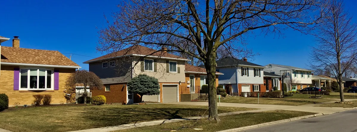 A street of 1950s and 1960s split-level and single-story houses with brick lower walls, lap siding above, shutters and attached garages, seen on a sunny early spring day