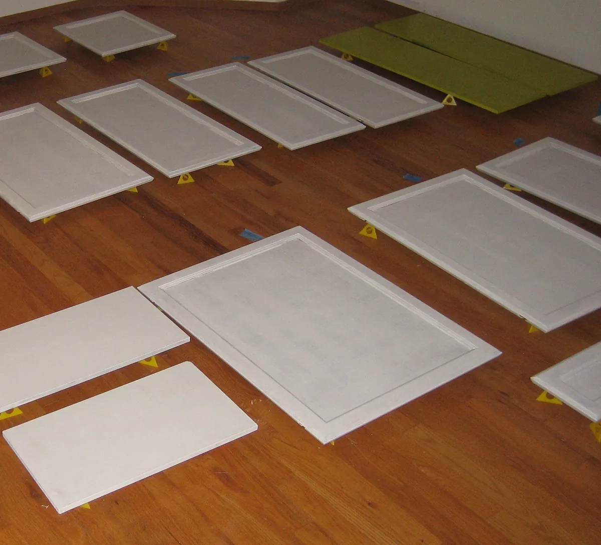 Primed white cabinet doors laid flat on yellow painter's pyramids across a wood floor, each door spaced apart, with a few lime green panels along the back wall