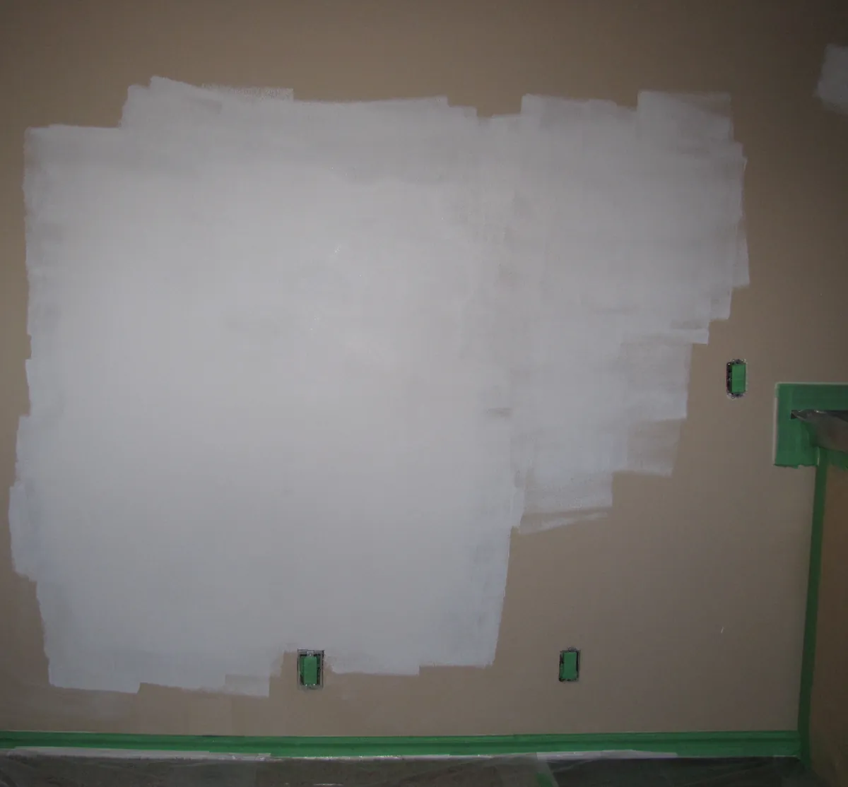 A tan living room wall partly covered in fresh white primer laid on in overlapping roller passes, with green painter's tape on the baseboard, door casing and outlets and plastic sheeting on the floor