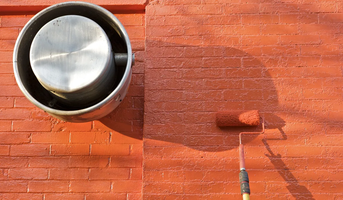 Paint roller on an extension pole resting against a freshly painted orange brick exterior wall in bright sun, beside a round metal vent
