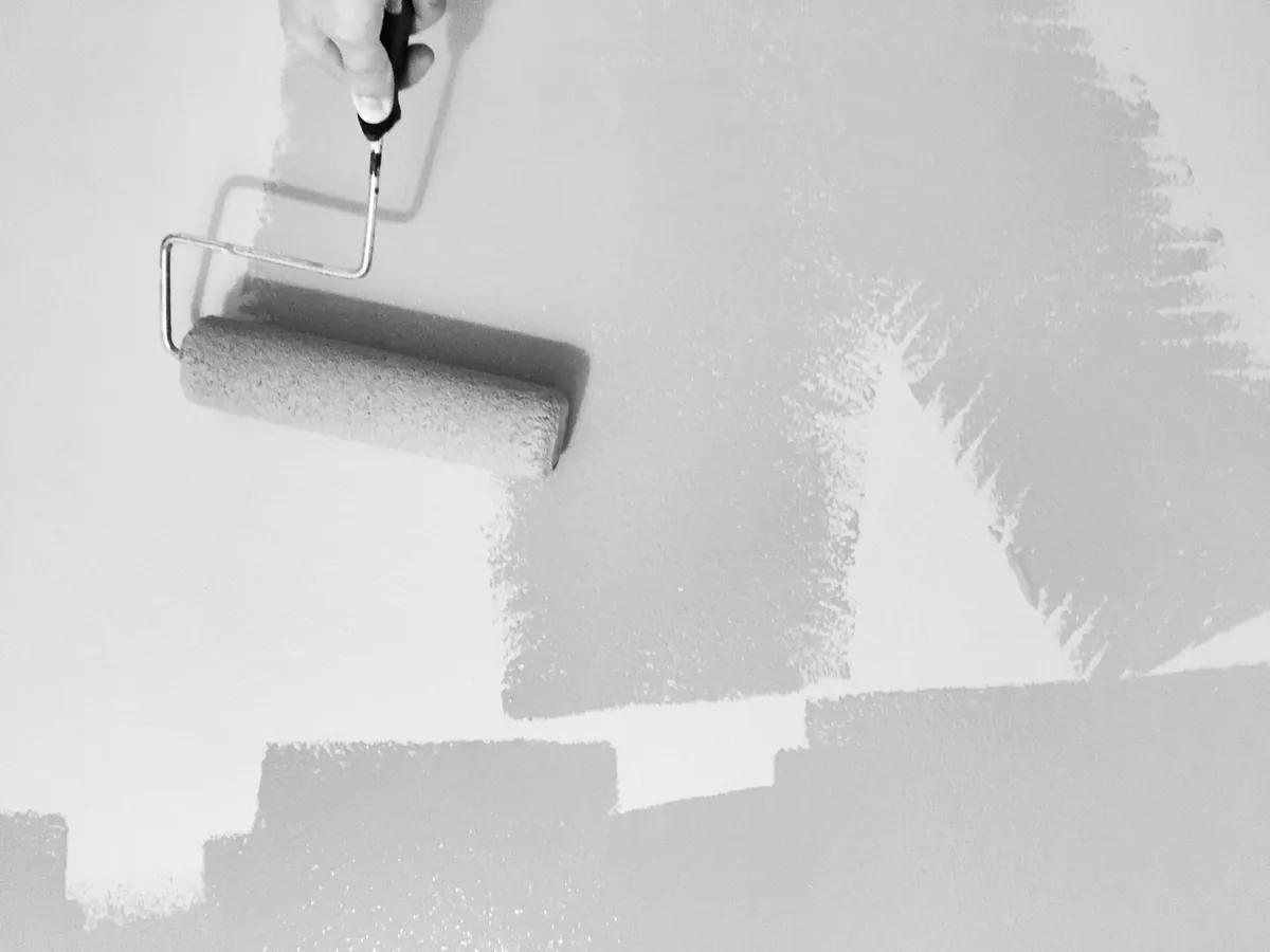 A hand rolling gray paint onto a white wall with a nap roller on a metal frame, the wall partly covered