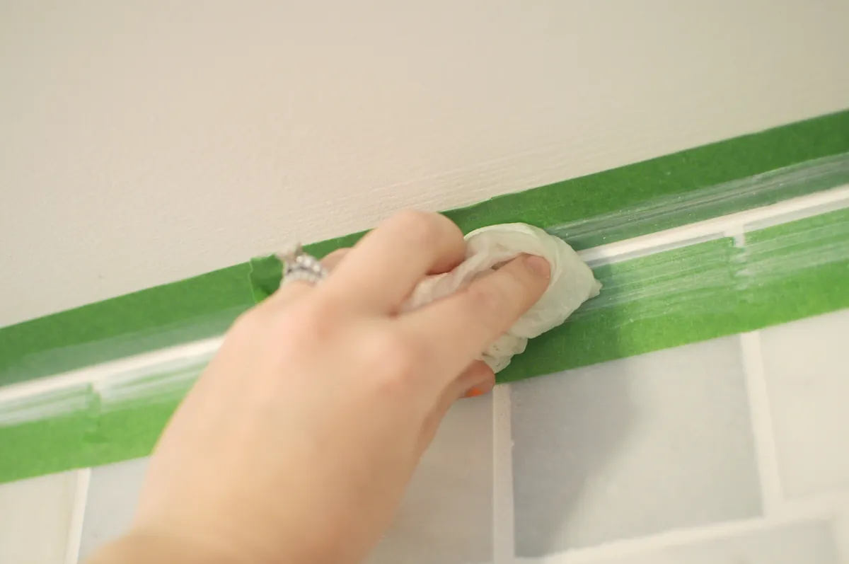 A hand smoothing a fresh bead of white caulk with a damp paper towel, between two strips of green painter's tape where a painted wall meets white tile