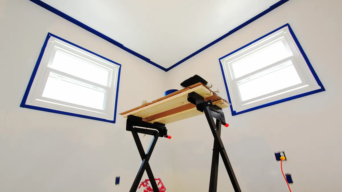 Bright room with white walls where blue painter's tape outlines two windows and the ceiling line, outlet covers removed, and a folding work table in the middle