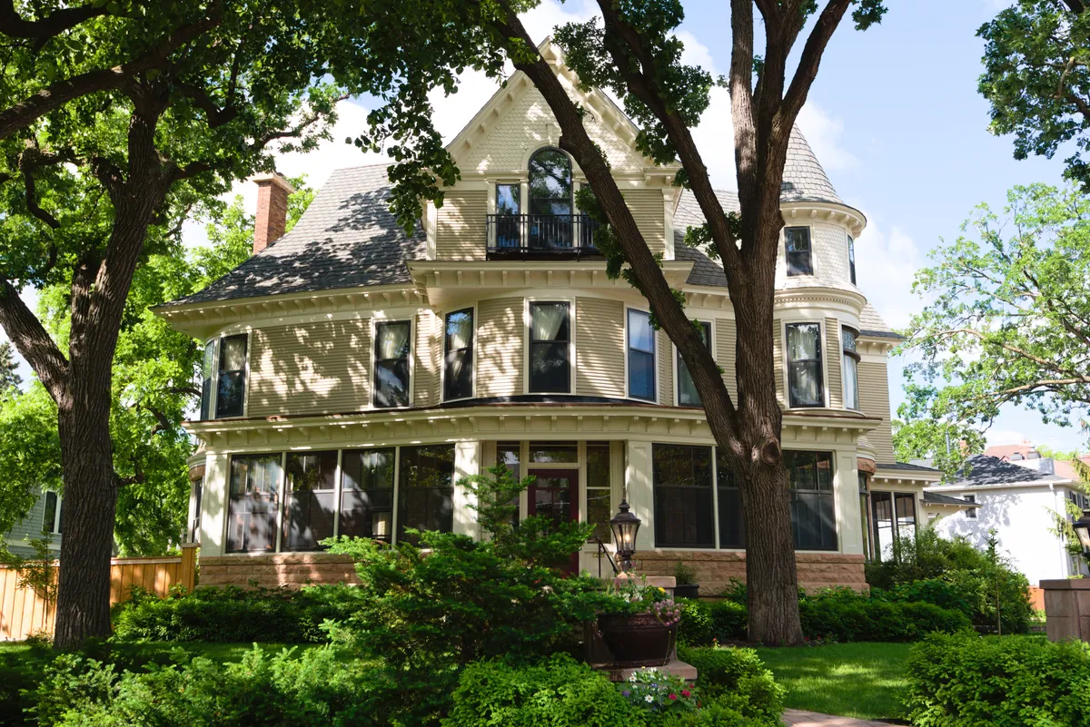 A large Victorian-era house in the Kenwood neighborhood of Minneapolis with cream painted lap siding, fish-scale shingles in the gable, a corner turret, dentil molding and a glassed-in front porch, shaded by mature trees