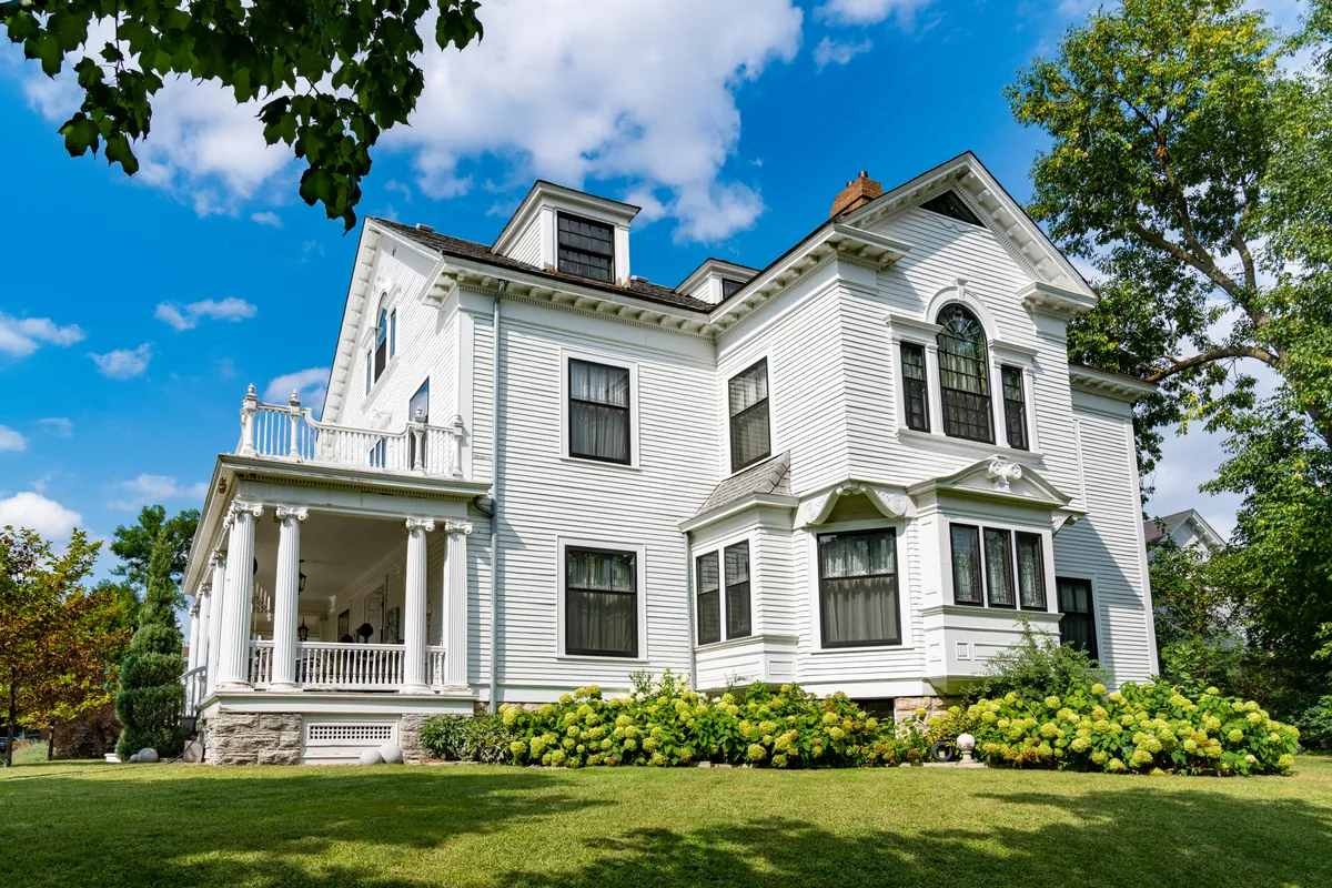 Large white clapboard house with white trim, a columned wraparound porch, black framed windows and hydrangeas along the foundation, under a blue summer sky