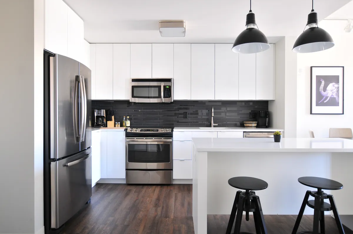 Bright white kitchen with flat white ceiling and walls, smooth white cabinet doors, a dark glossy tile backsplash, stainless appliances, black pendant lights and two black stools at a white island